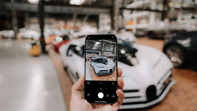 Durch eine Smartphone-Kamera ist ein weißes Sportauto zu sehen.