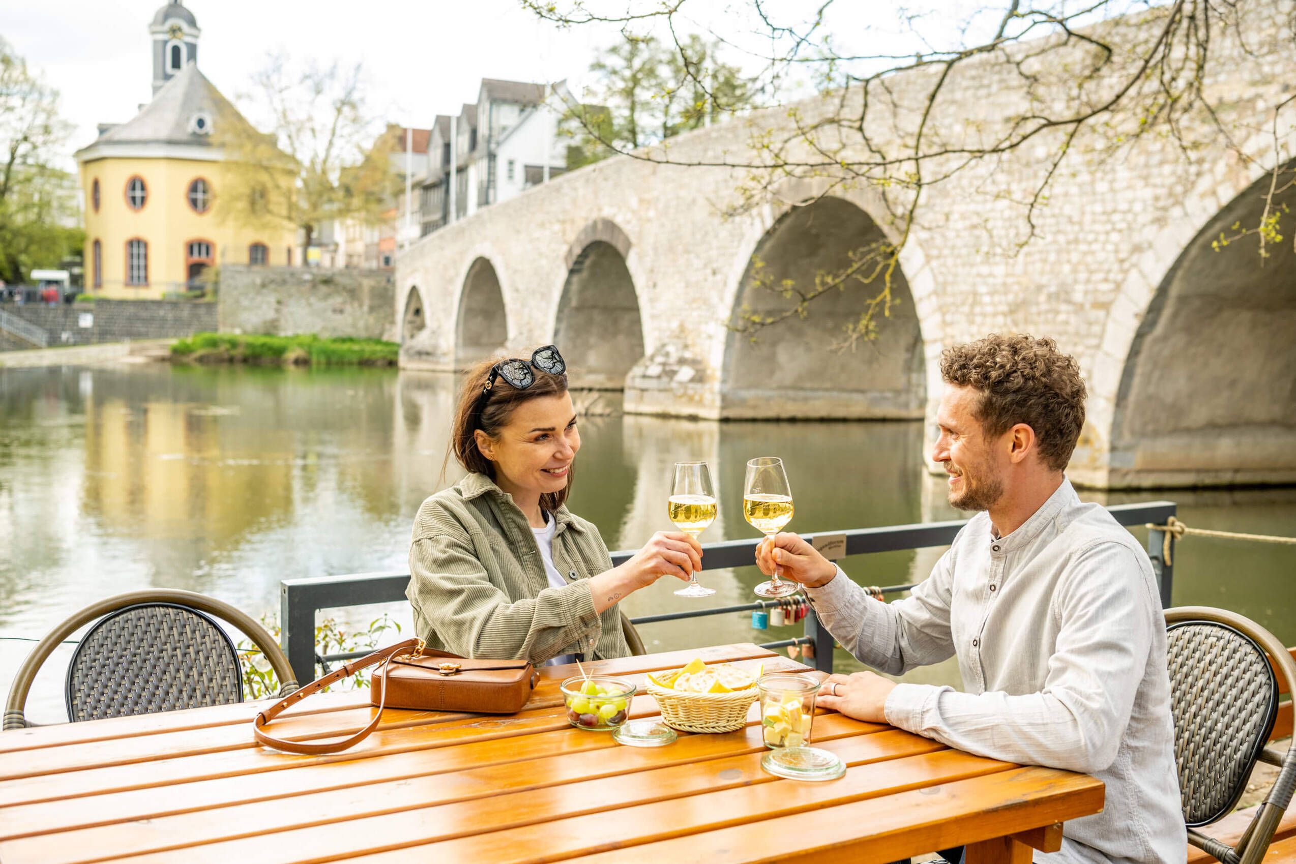 Zwei Personen prosten sich am Ufer der Lahn mit Weingläsern zu.
